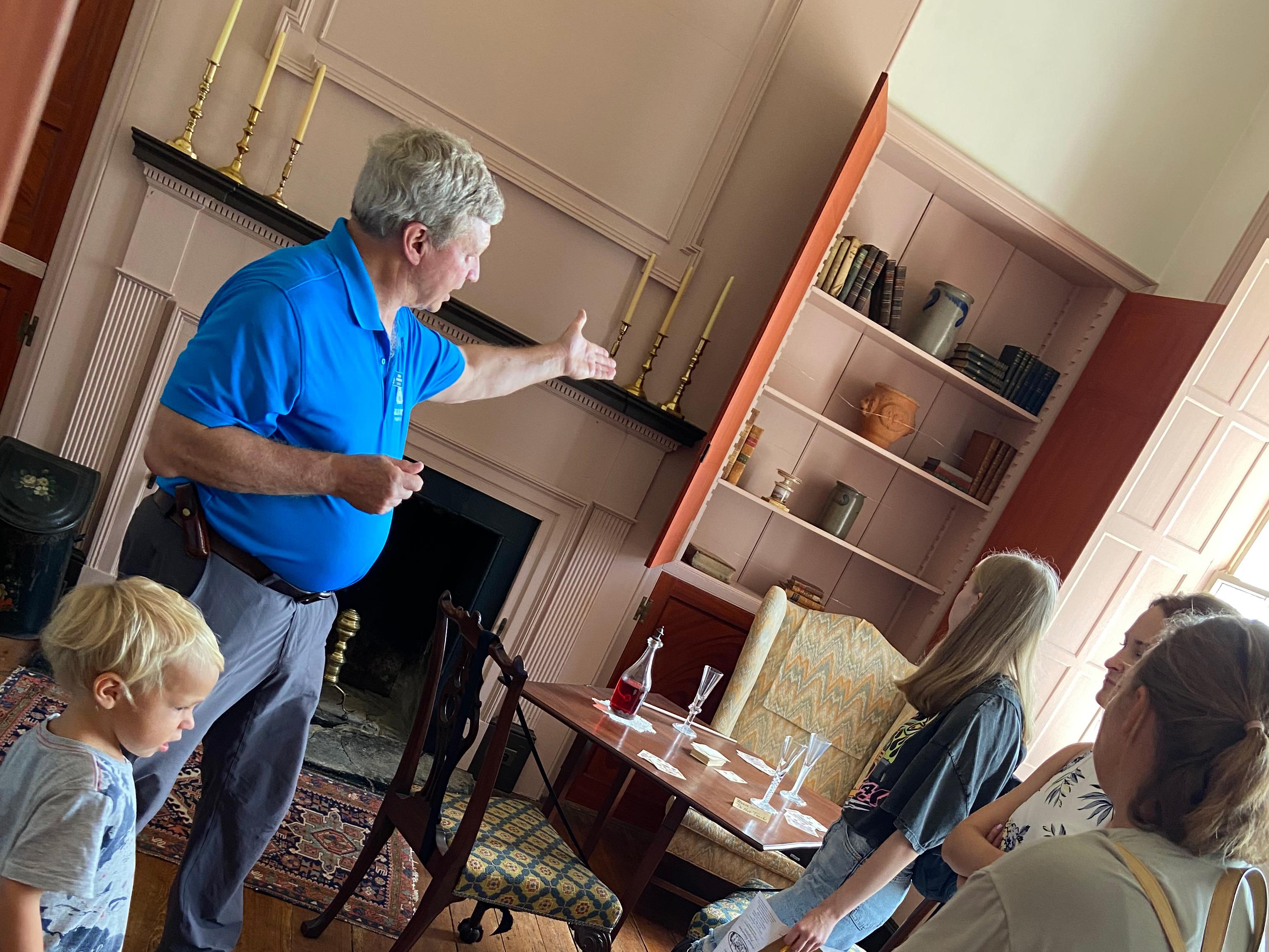 A man in a blue shirt leading a tour group gestures to items in a 1700s mansion.