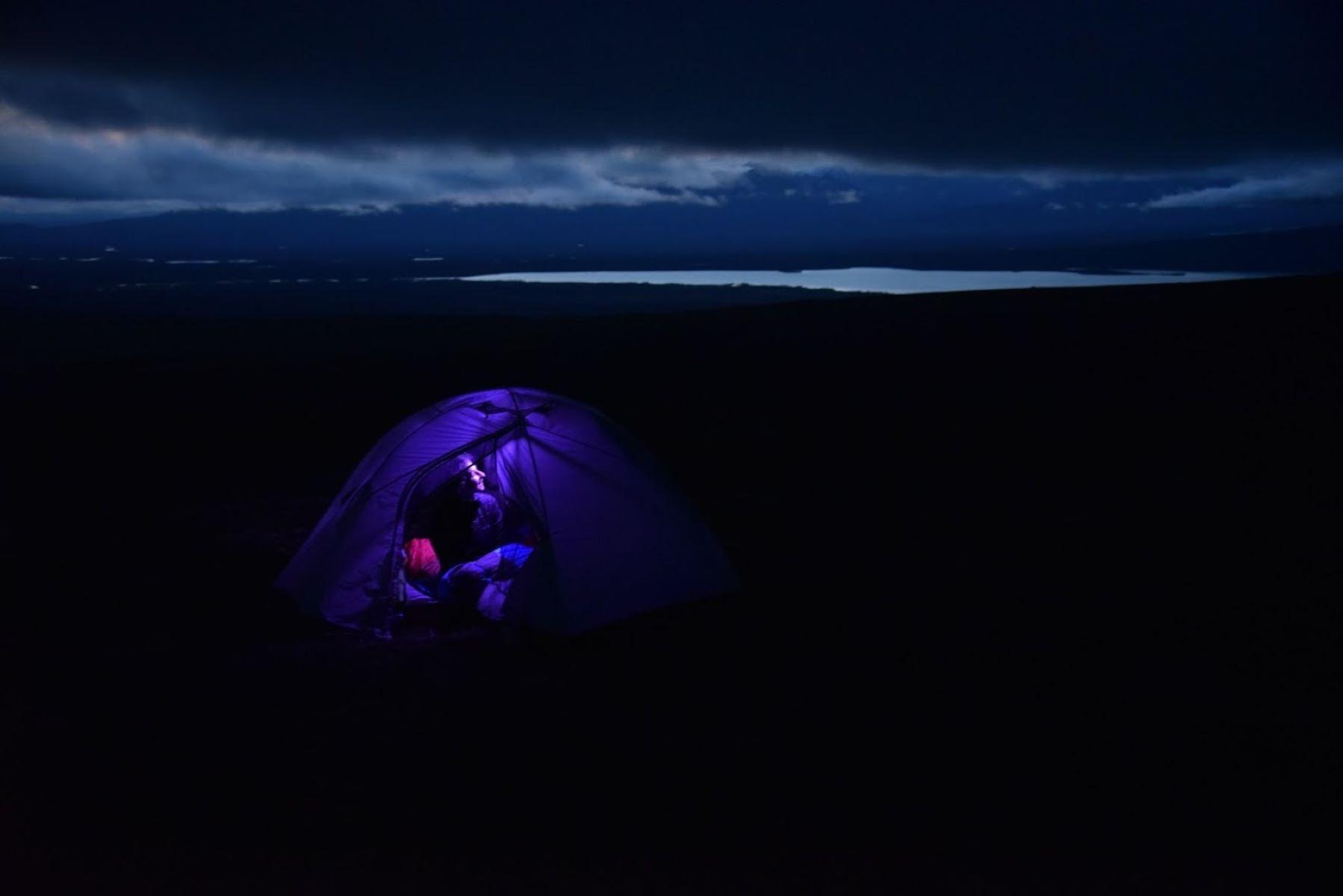 A night time scene of a woman in a tent with her headlamp glowing up the walls.