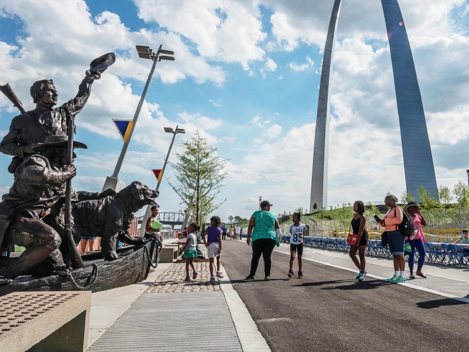 a group of people standing along a trail next to the Captain's Return statue in front of the Arch