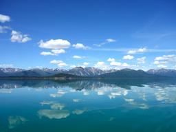 Photo of blue sky with fluffy white clouds reflect in calm lake with mountains in the background.