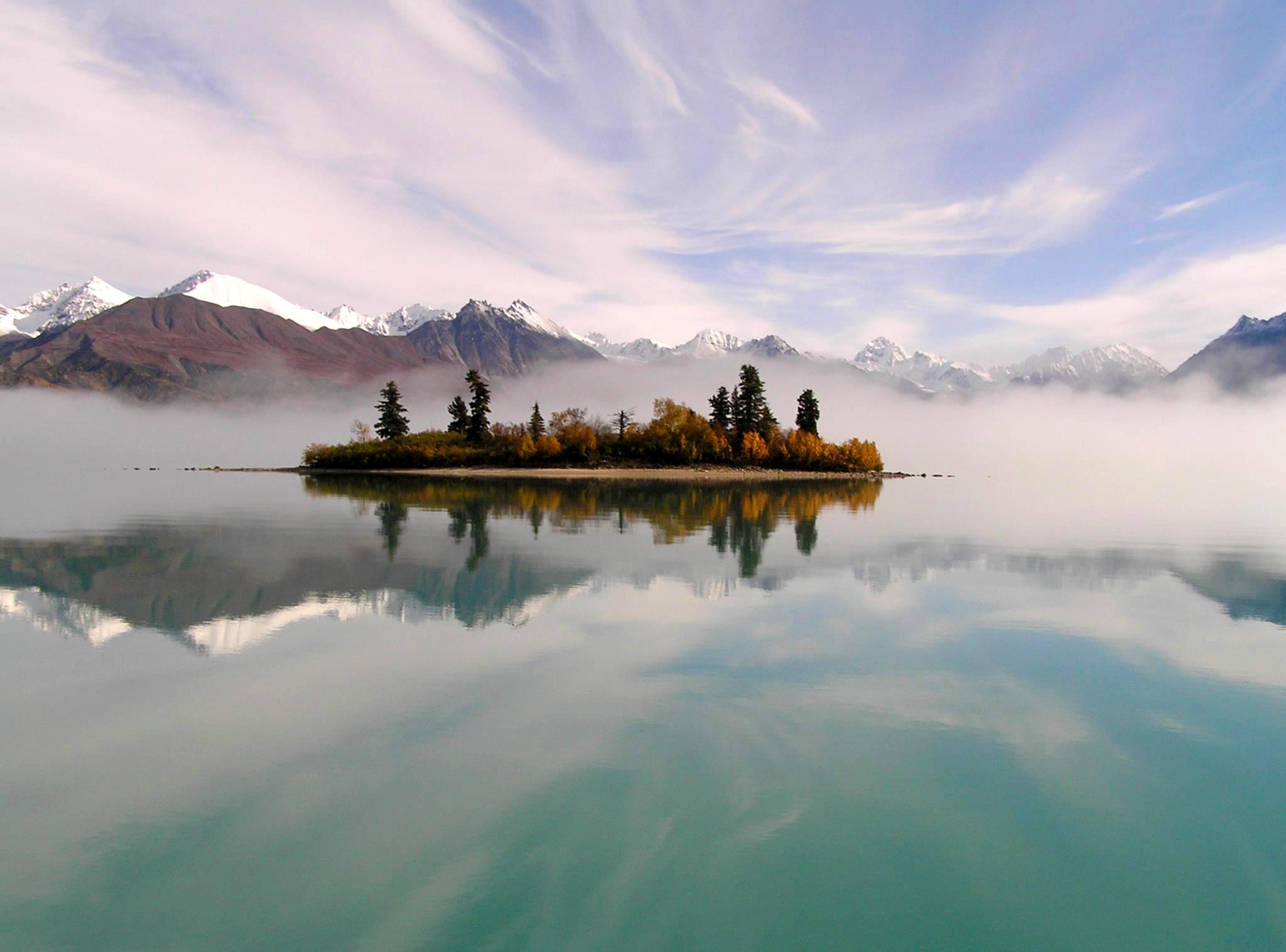 A colorful sky, an island with spruce trees, and mountains in the background reflect in a calm lake