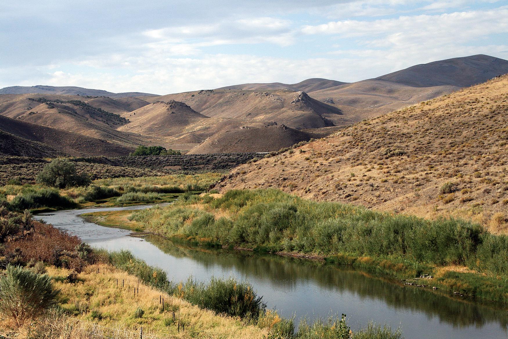 A still creek winds through brown rolling hills.