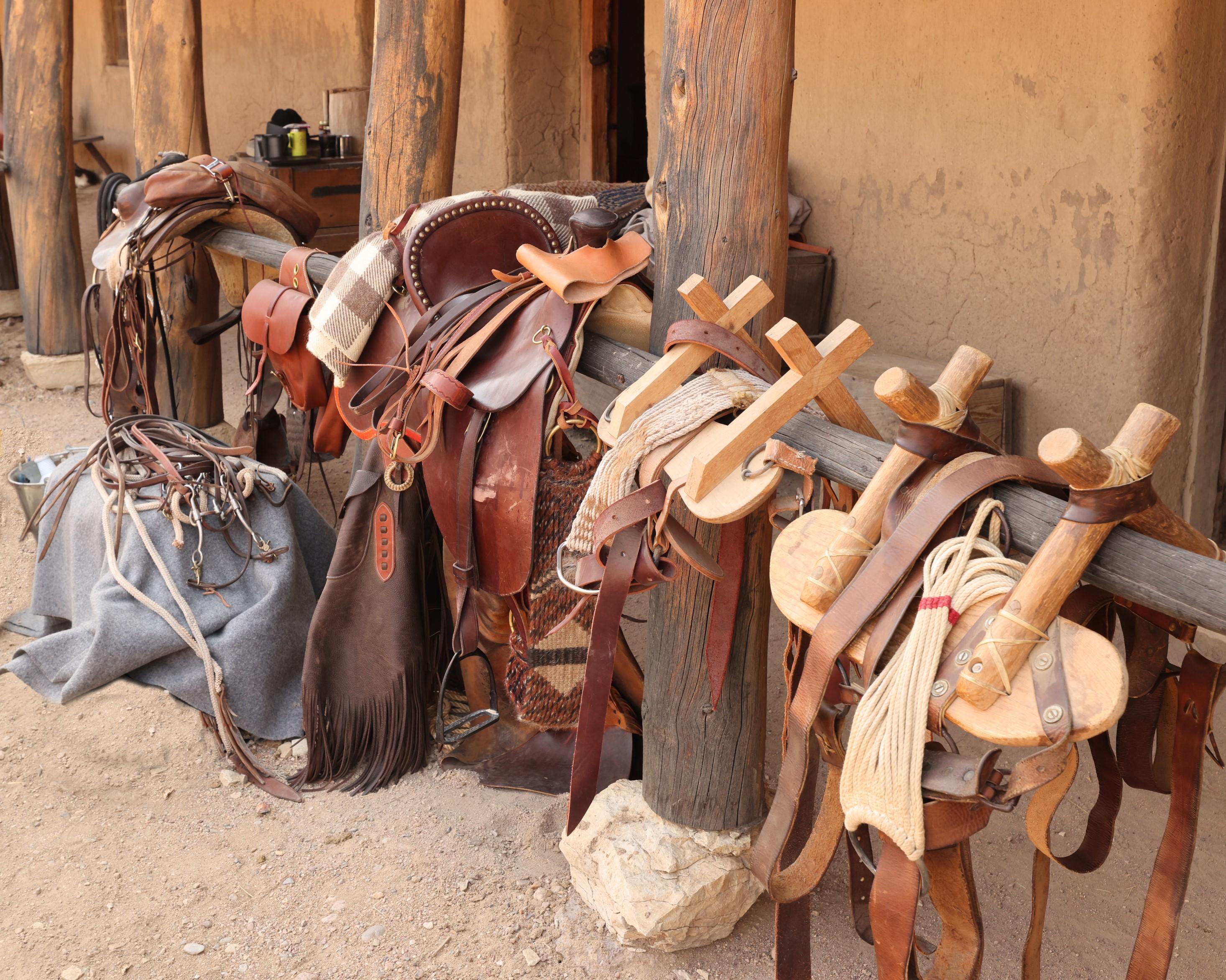 A row of saddles are arrayed along a wooden beam near an adobe building